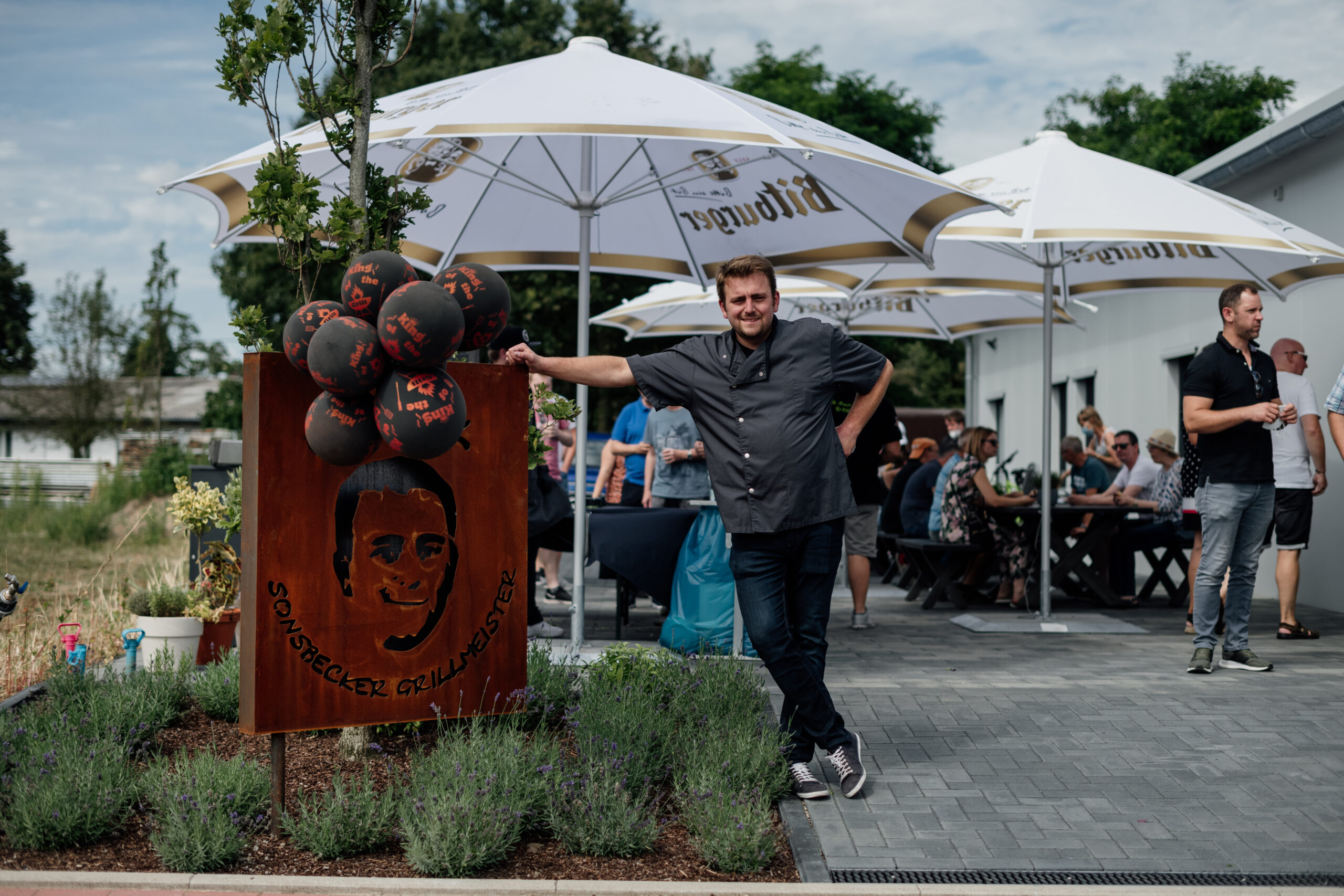 Original Sonsbecker Grillmeister Christian Holz steht stolz an sein Schild angelehnt, im Hintergrund sitzen Leute und genießen den Sommertag beim Essen